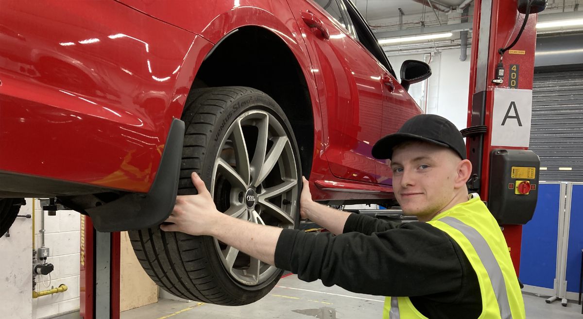 Young man wearing black top and baseball cap and high viz jacket holding car wheel attached to elevated vehicle in garage.