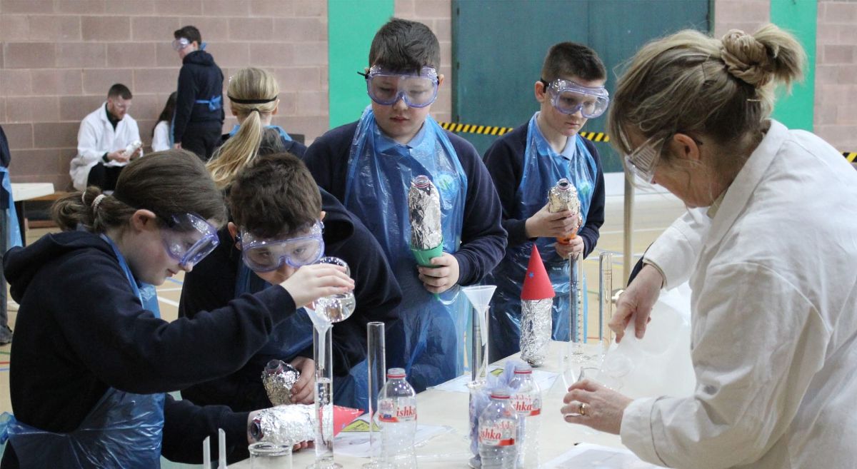 Four children in school uniforms, wearing plastic aprons and face shields, pouring liquids into containers with woman in white lab coat.