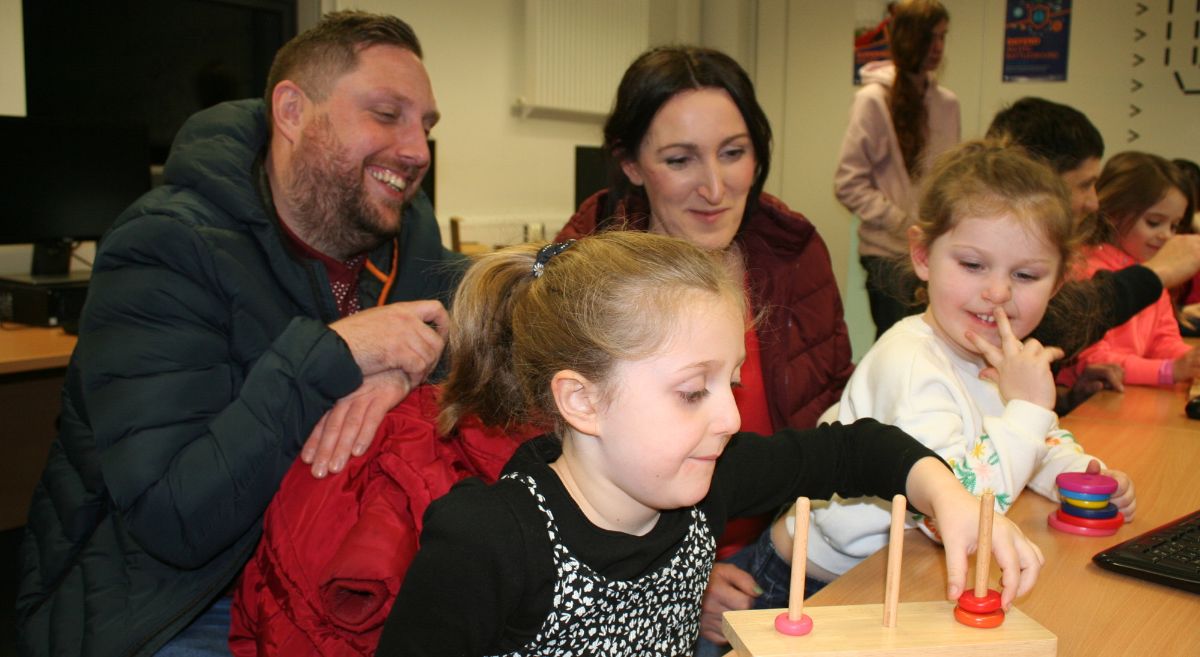 Two parents watching over the shoulder of two children doing a stacking puzzle.