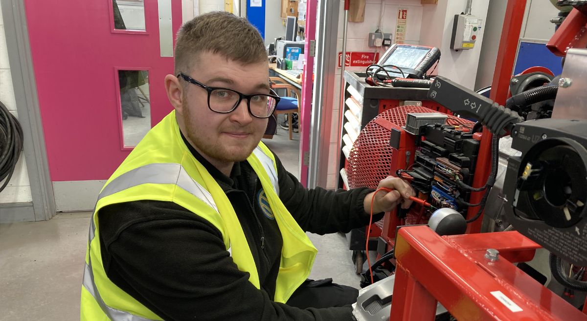 Young man wearing black top and high viz jacket working at engine in training garage.