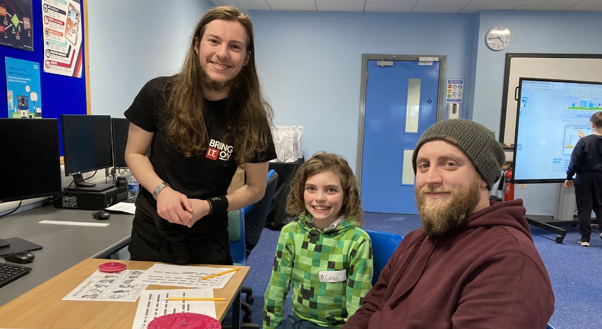 Student in a black teeshirt with a parent and child working at a puzzle in a computer class rooom.