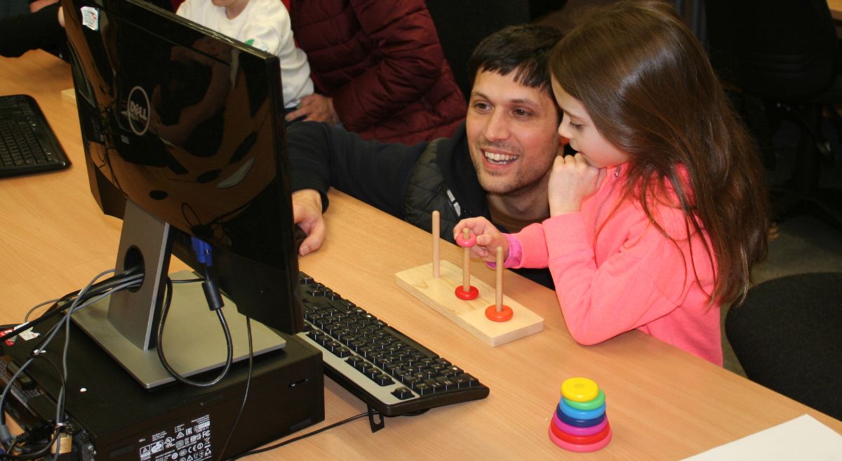 A male parent beside a child with long dark hair and pick top working at a desktop computer with a stacking puzzle in front of the keyboard.