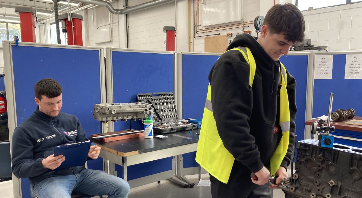 Man sitting with clip board  with another wearing back top and high viz works at engine part .