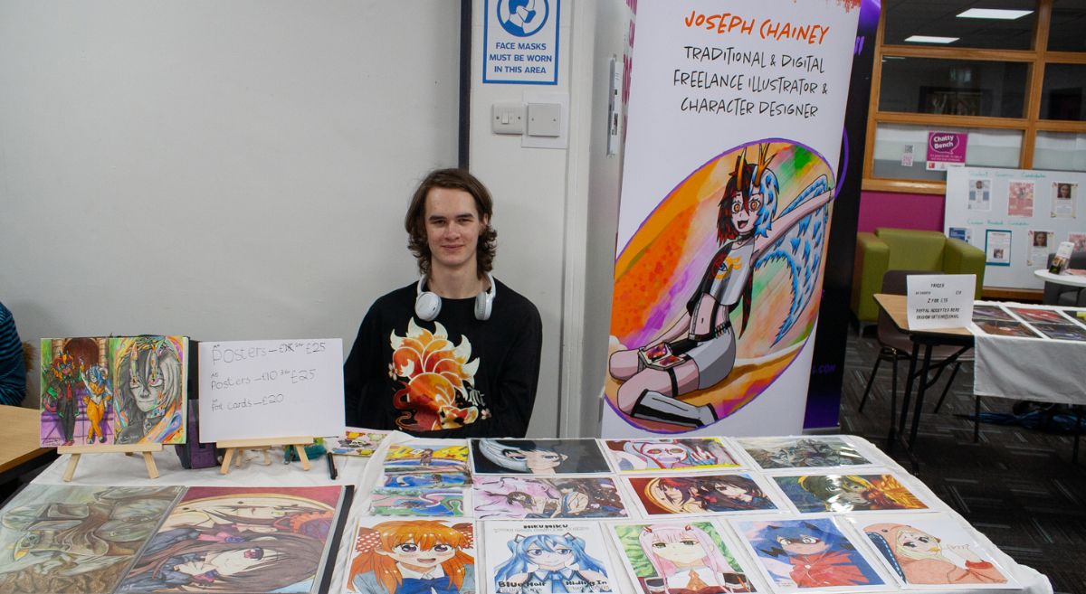 A person sat behind a table selling illustrations, a roll up banner in the background