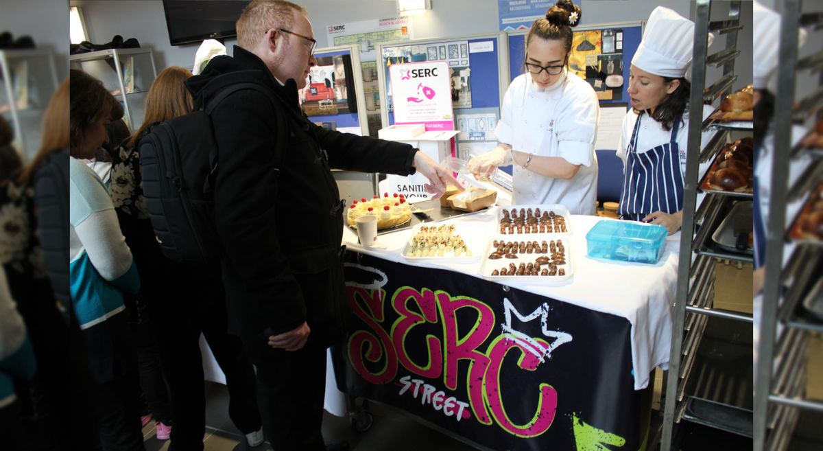 a person enquiring about food products, two members of a student company behind the table advising/selling products