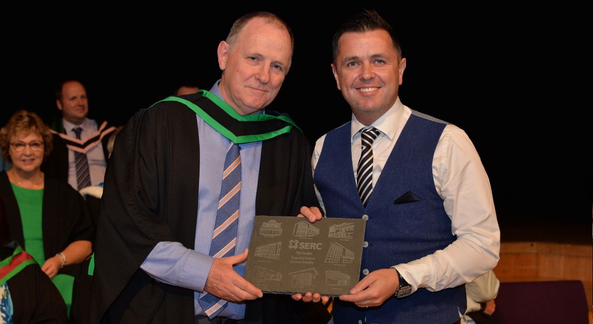 A man in a graduation gown presents award to man in white shirt, stripped tie and wasitcoat