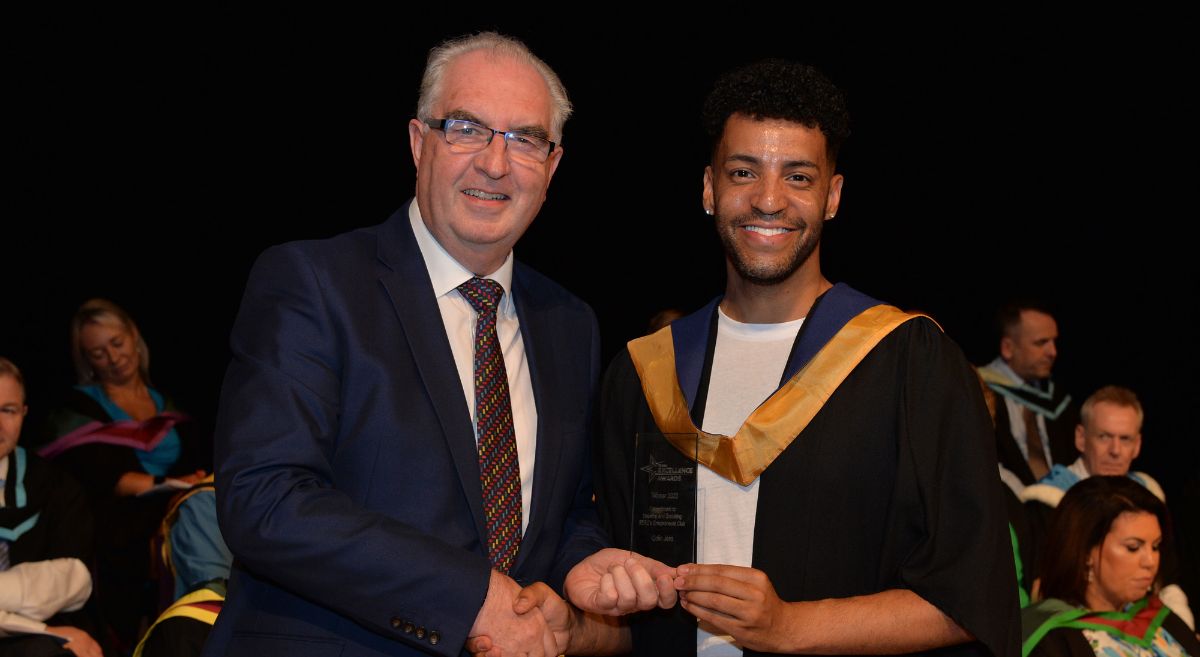 A man in a dark suit receiving award from man in graduation gown