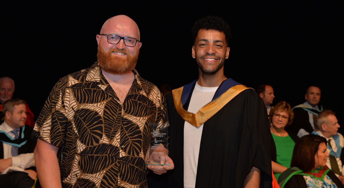 a man in a patterned shirt with red beard and glasses receiving an award from a man in graduation gown