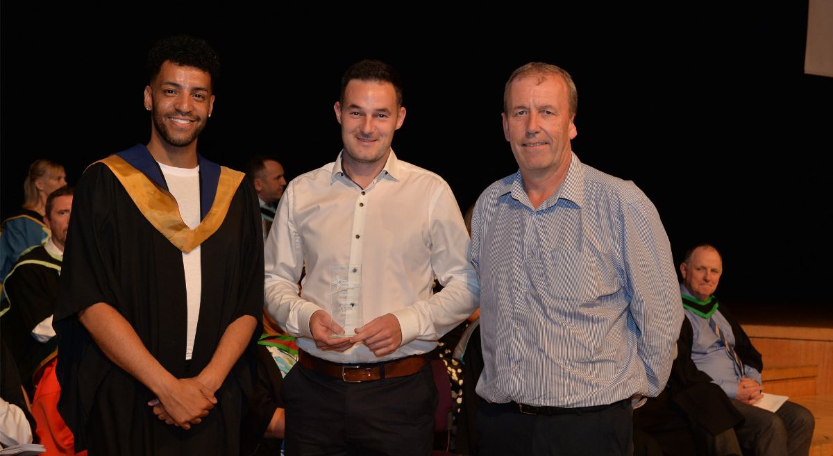 A man in a graduation gown presents award to man in white shirt with a man in a stripped shirt