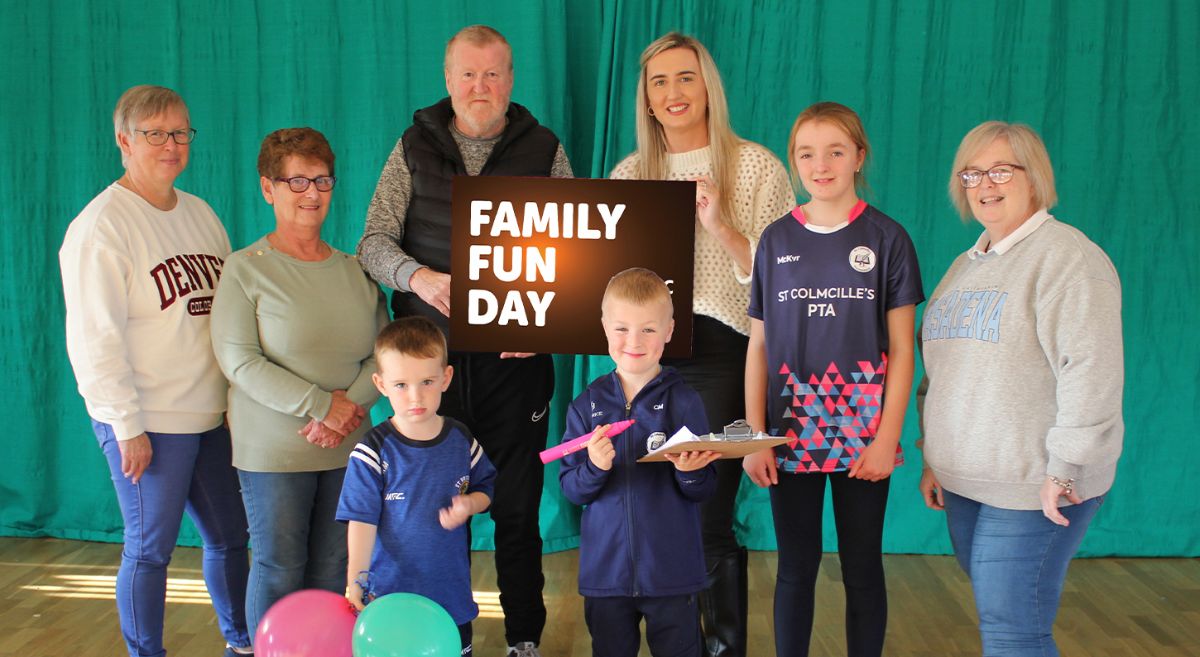 Five adults and three children pose with a Family Fun Day Sign,  Two children hold balloons and clipboard and giant pen.