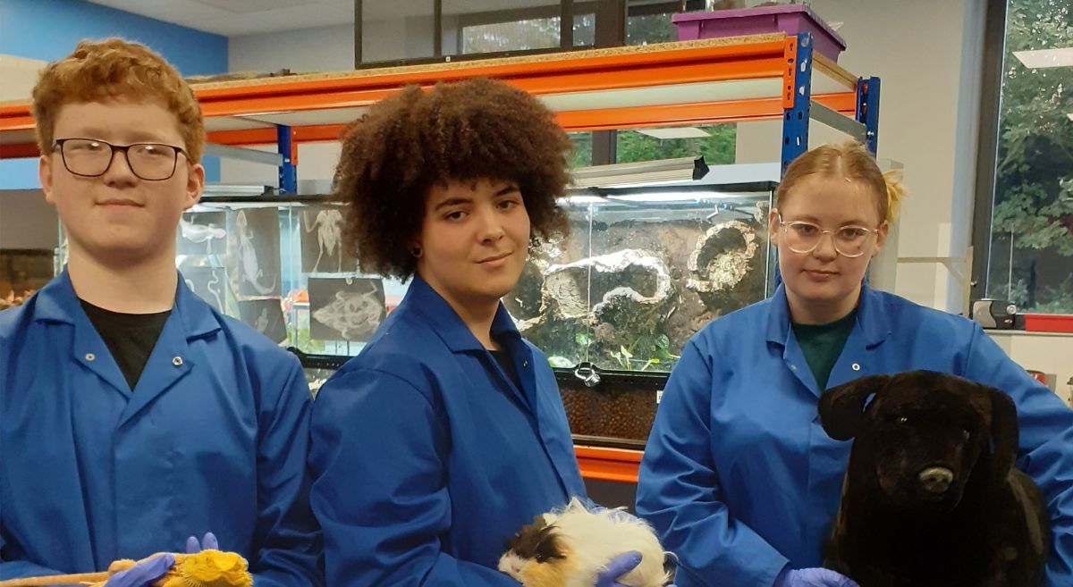 Three students, two with live animals and one with model dog, all wearing blue lab coats in animal house.