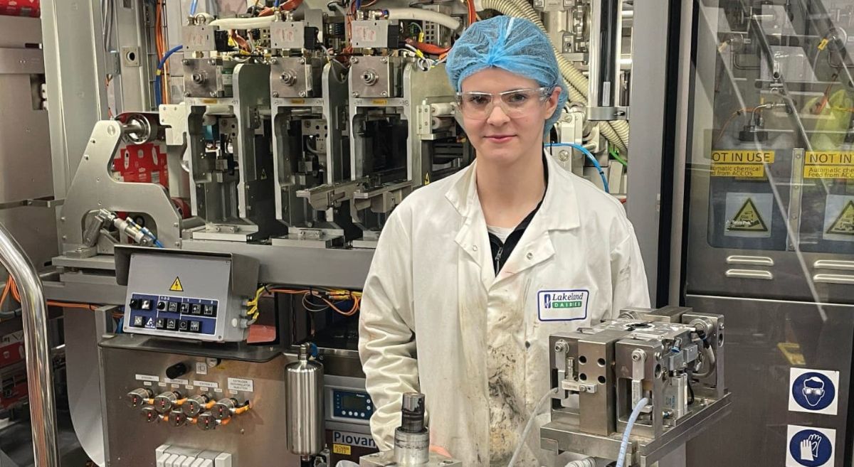 Female wearing white coat and blue hairnet in factory setting.