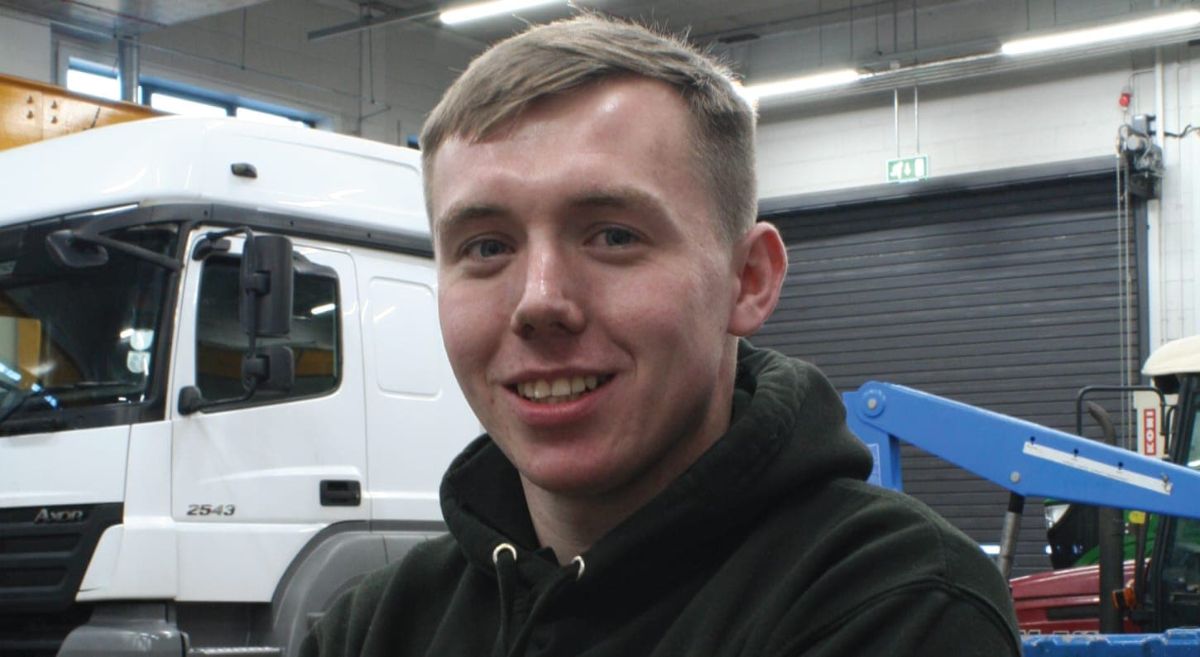 Young male smiling to camera with lorry in background in vehicle workshop.